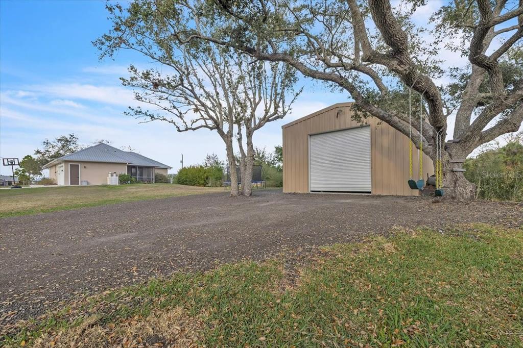 A detached metal garage sits on a gravel driveway near a house and large trees — 8429 SW RIVERVIEW OAKS TERRACE, ARCADIA, FL
