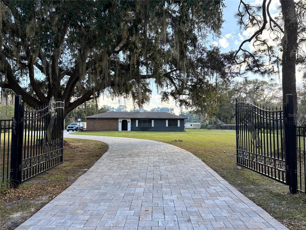 A paved driveway leads through an open gate toward a single-story house under large trees — 1800 GRANADA BOULEVARD, KISSIMMEE, FL