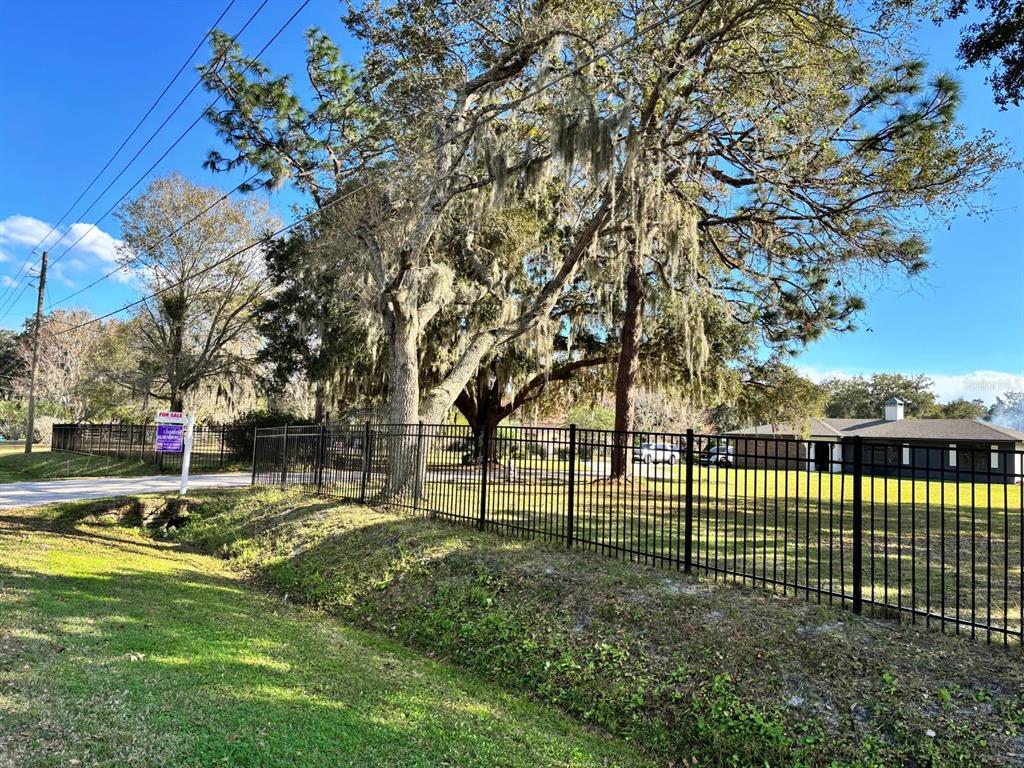 A black metal fence encloses a grassy yard with large trees under a blue sky — 1800 GRANADA BOULEVARD, KISSIMMEE, FL