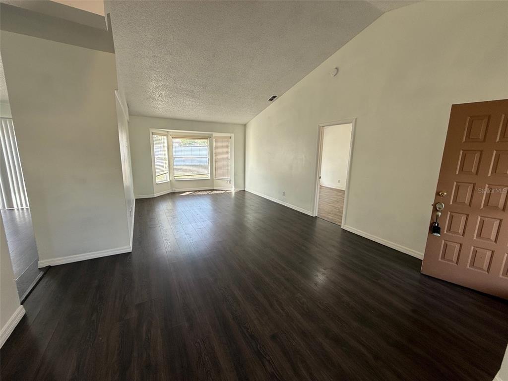 An empty living room features dark wood-look flooring, a vaulted ceiling, and a bay window — 13102 LINDEN DRIVE, SPRING HILL, FL