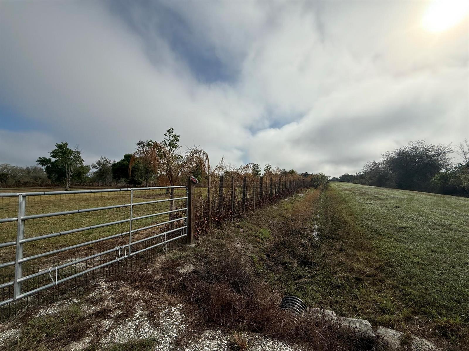 A metal gate opens to a fenced pasture beside a grassy embankment under a cloudy sky — 17231 NW 268th Street, Okeechobee FL 34972, OKEECHOBEE, FL