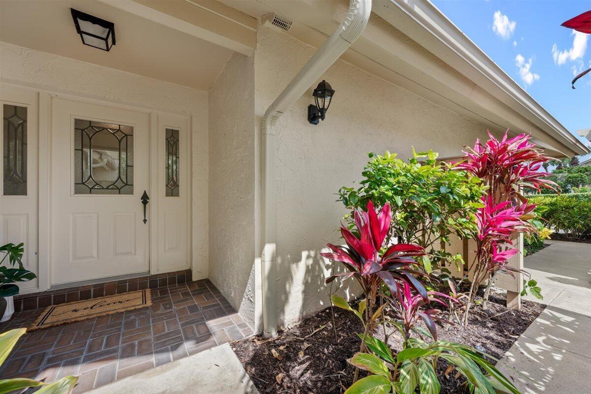 A white front door with a glass insert sits beside a garden with tropical plants — 17138 Waterbend Drive # 117, Jupiter FL 33477, JUPITER, FL