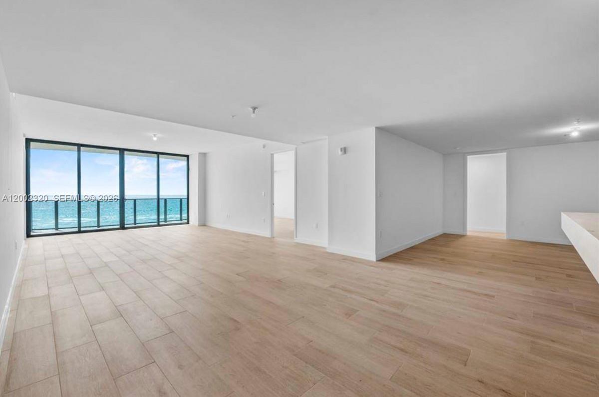 An empty, modern living room features light wood-look flooring, white walls, and floor-to-ceiling windows — 900 N Ocean Blvd # 1202, Pompano Beach FL 33062, POMPANO BEACH, FL