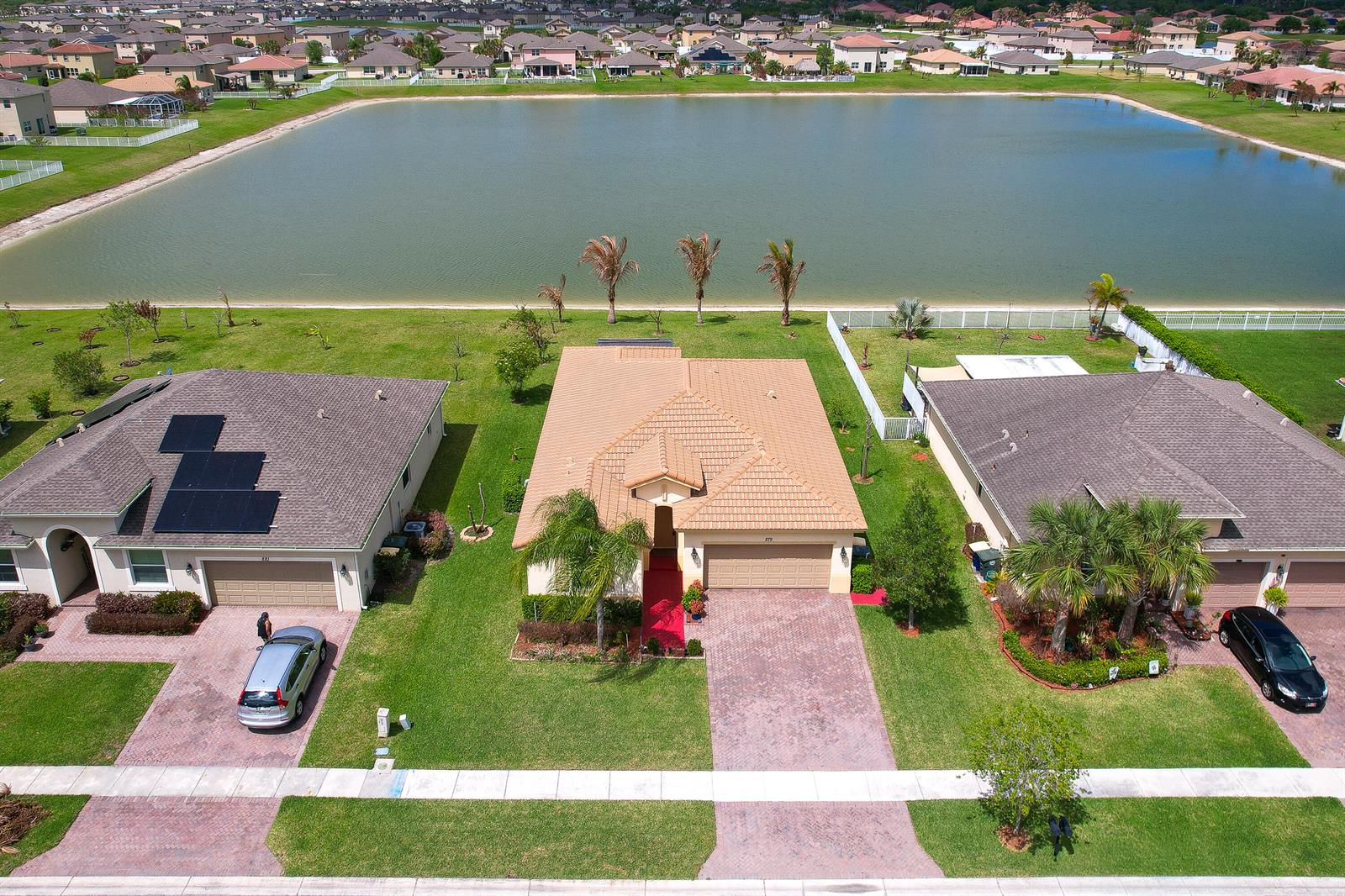 An aerial view shows three suburban houses with tiled roofs situated along a large lake — 879 Bent Creek Drive, Fort Pierce FL 34947, FORT PIERCE, FL
