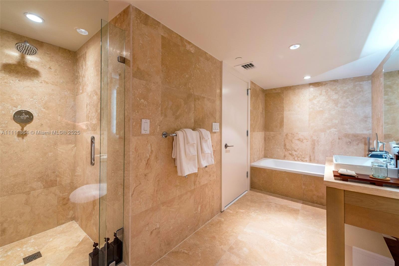This bathroom features beige stone tile walls and flooring, a glass-enclosed shower, and a bathtub — 102 24th St # 906, Miami Beach FL 33139, MIAMI BEACH, FL