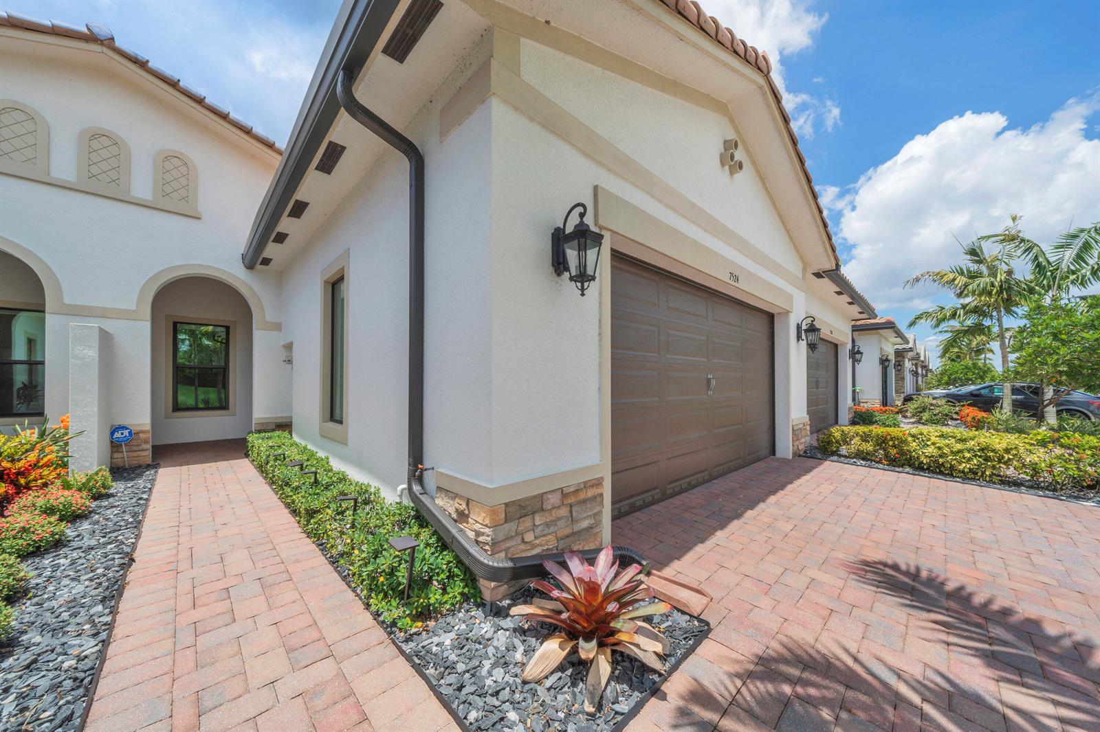 A stucco house exterior features a brown garage door, brick driveway, and landscaped garden beds — 7524 N Blue Spring Drive, Parkland FL 33067, PARKLAND, FL