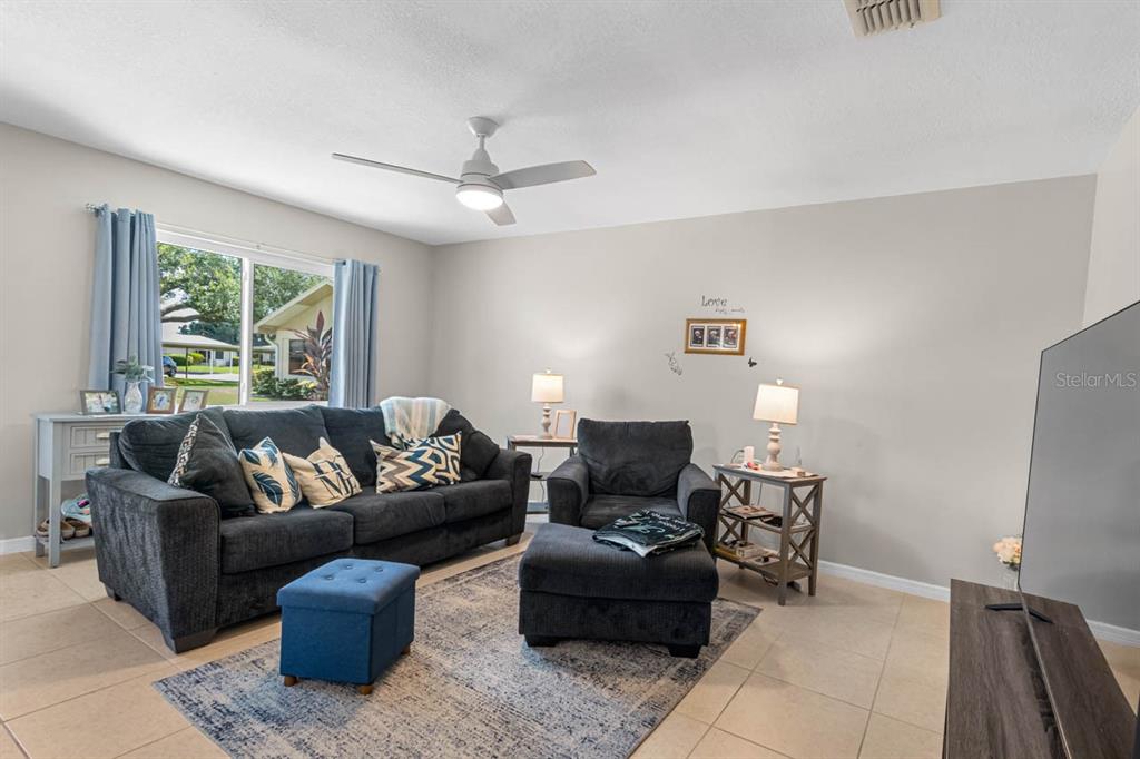 A living room features a dark sofa, armchair, ottoman, area rug, and a ceiling fan — 1906 CANTERBURY LANE UNIT 26, SUN CITY CENTER, FL