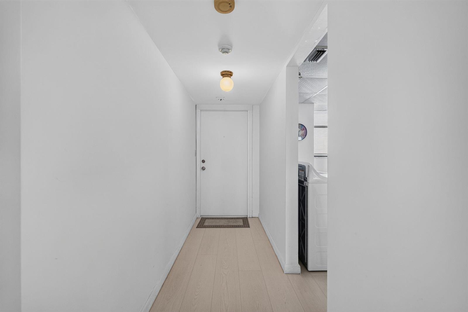 A hallway with light wood-look flooring leads to a white door and an adjacent laundry area — 9900 S Ocean Drive # 1505, Jensen Beach FL 34957, JENSEN BEACH, FL