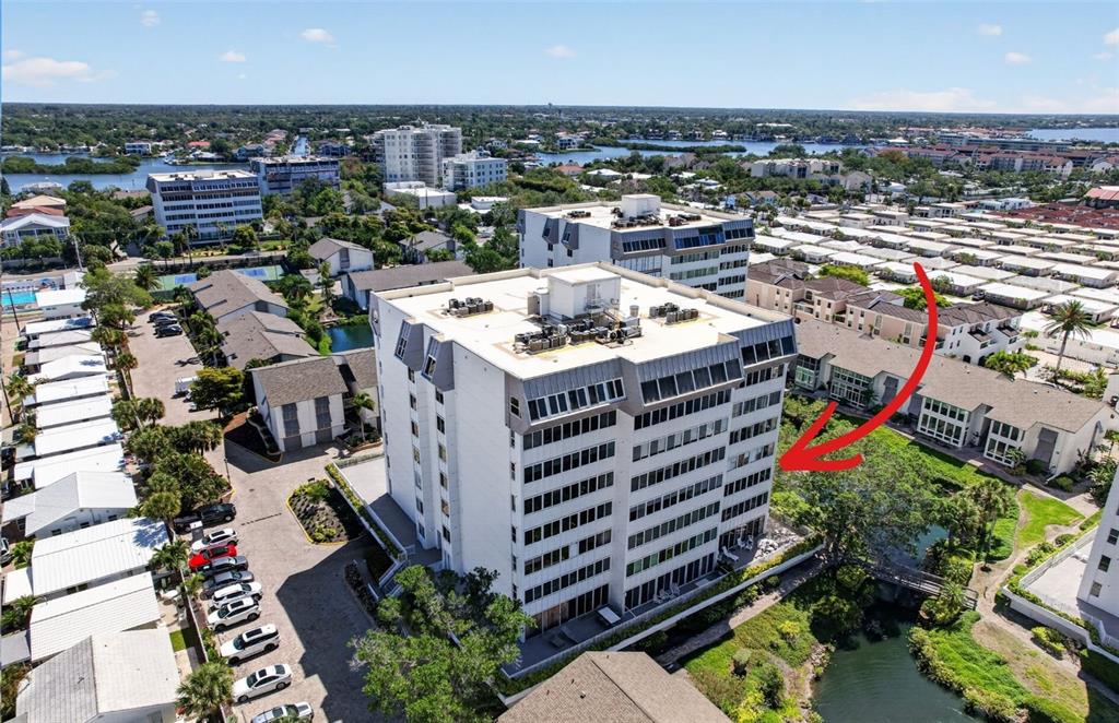 An aerial view shows a multi-story white apartment building with a red arrow pointing toward it — 1080 W PEPPERTREE LANE UNIT 305A, SARASOTA, FL