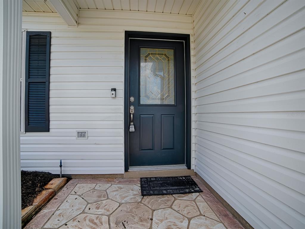 A dark front door with a glass insert sits on a stone porch with white siding — 1611 ALHAMBRA WAY, THE VILLAGES, FL