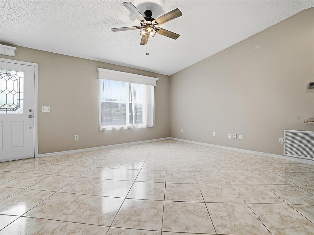 An empty room features beige walls, light-colored tile flooring, a ceiling fan, and a front door — 1611 ALHAMBRA WAY, THE VILLAGES, FL