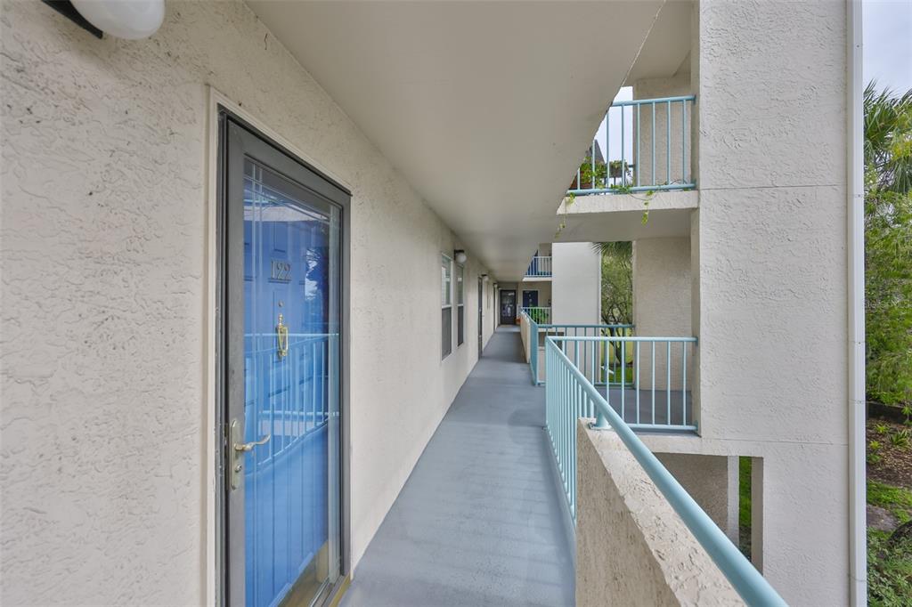 An outdoor apartment walkway features a blue front door, light-colored stucco walls, and railings — 5567 SEA FOREST DRIVE UNIT 122, NEW PORT RICHEY, FL