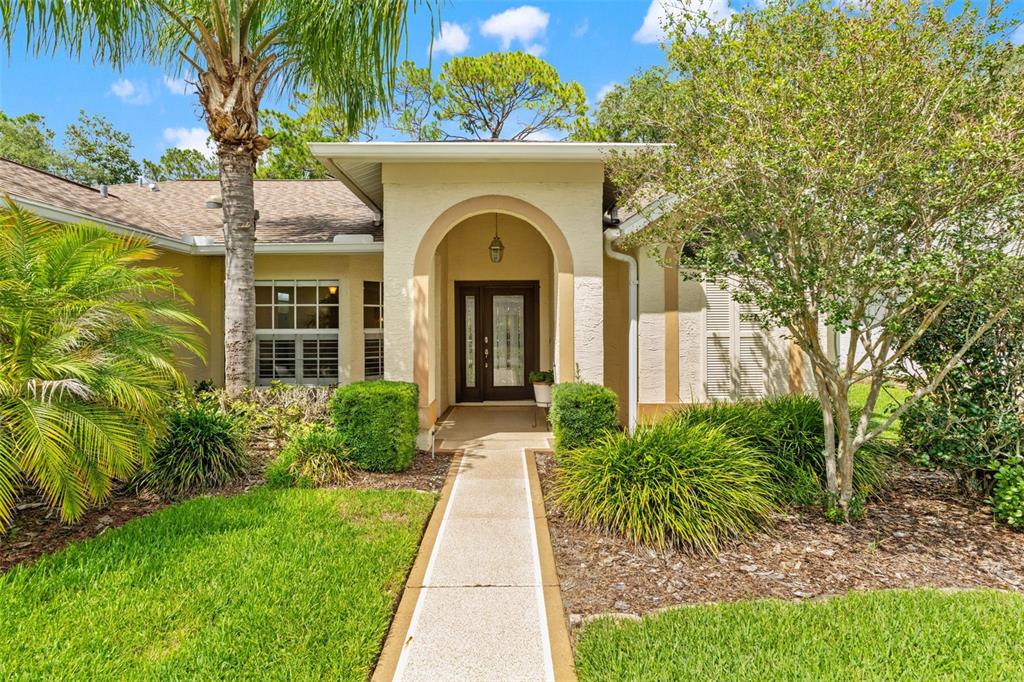A beige house exterior features an arched entryway, a paved walkway, and surrounding landscaping — 2593 CRYSTAL LAKE DRIVE, SPRING HILL, FL