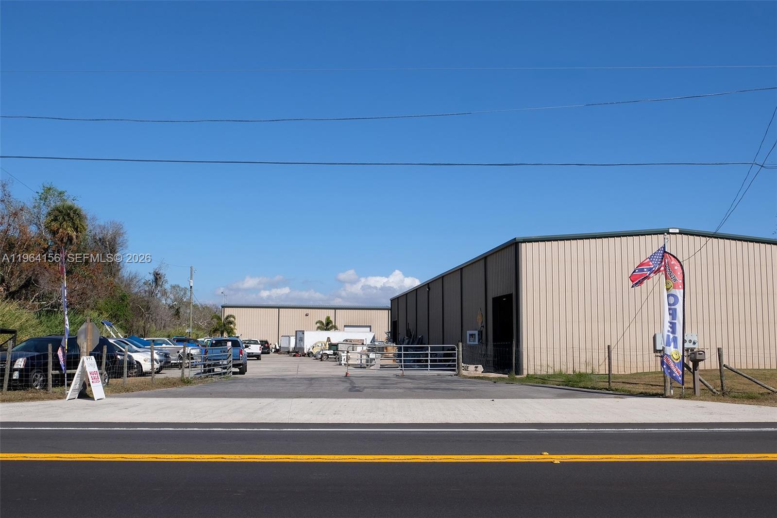 A paved lot features two metal warehouse buildings and several parked vehicles under a blue sky — 5271 US HWY 441 SE, Okeechobee FL 34974, OKEECHOBEE, FL