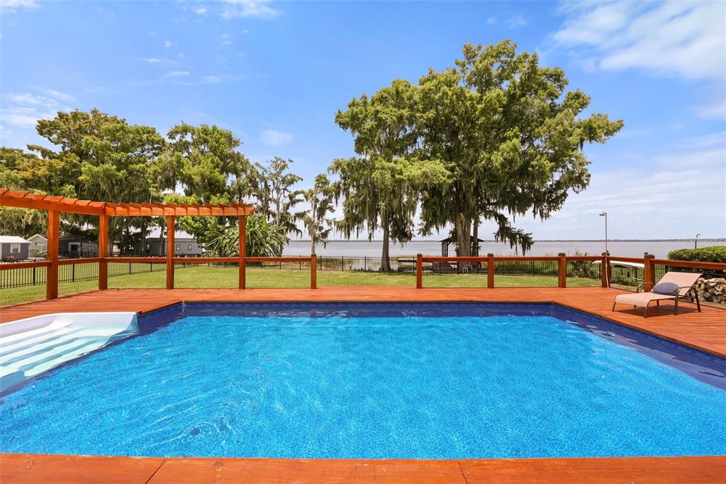 A rectangular swimming pool sits on a wooden deck overlooking a lake and large trees — 32749 LAKE EUSTIS DRIVE, TAVARES, FL