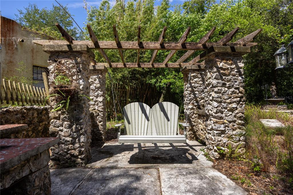 A stone pergola with a hanging bench swing sits on a concrete patio outdoors — 720 NE BOULEVARD, GAINESVILLE, FL