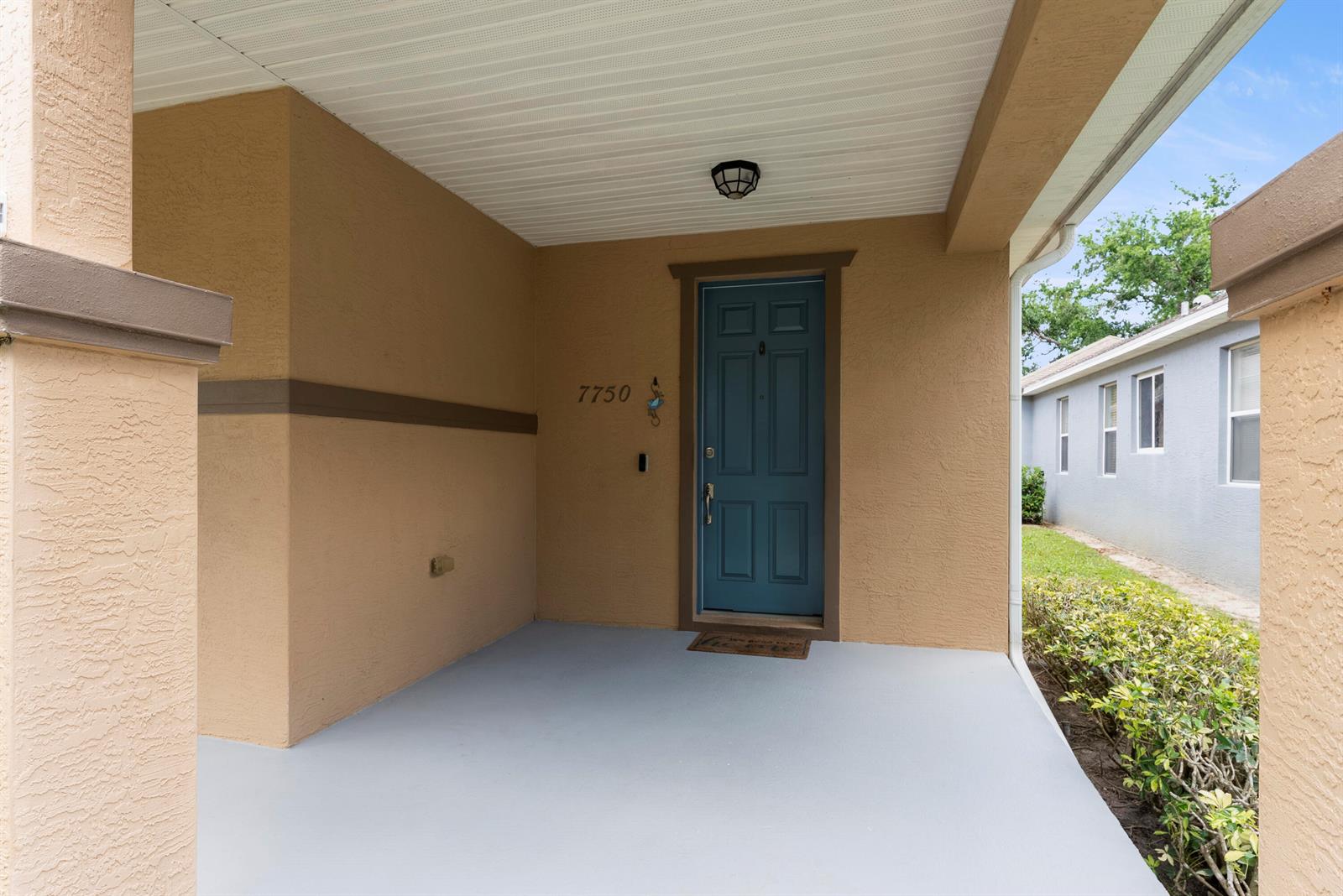 A covered front porch features a blue door, tan stucco walls, and a gray floor — 7750 15th Street, Vero Beach FL 32966, VERO BEACH, FL
