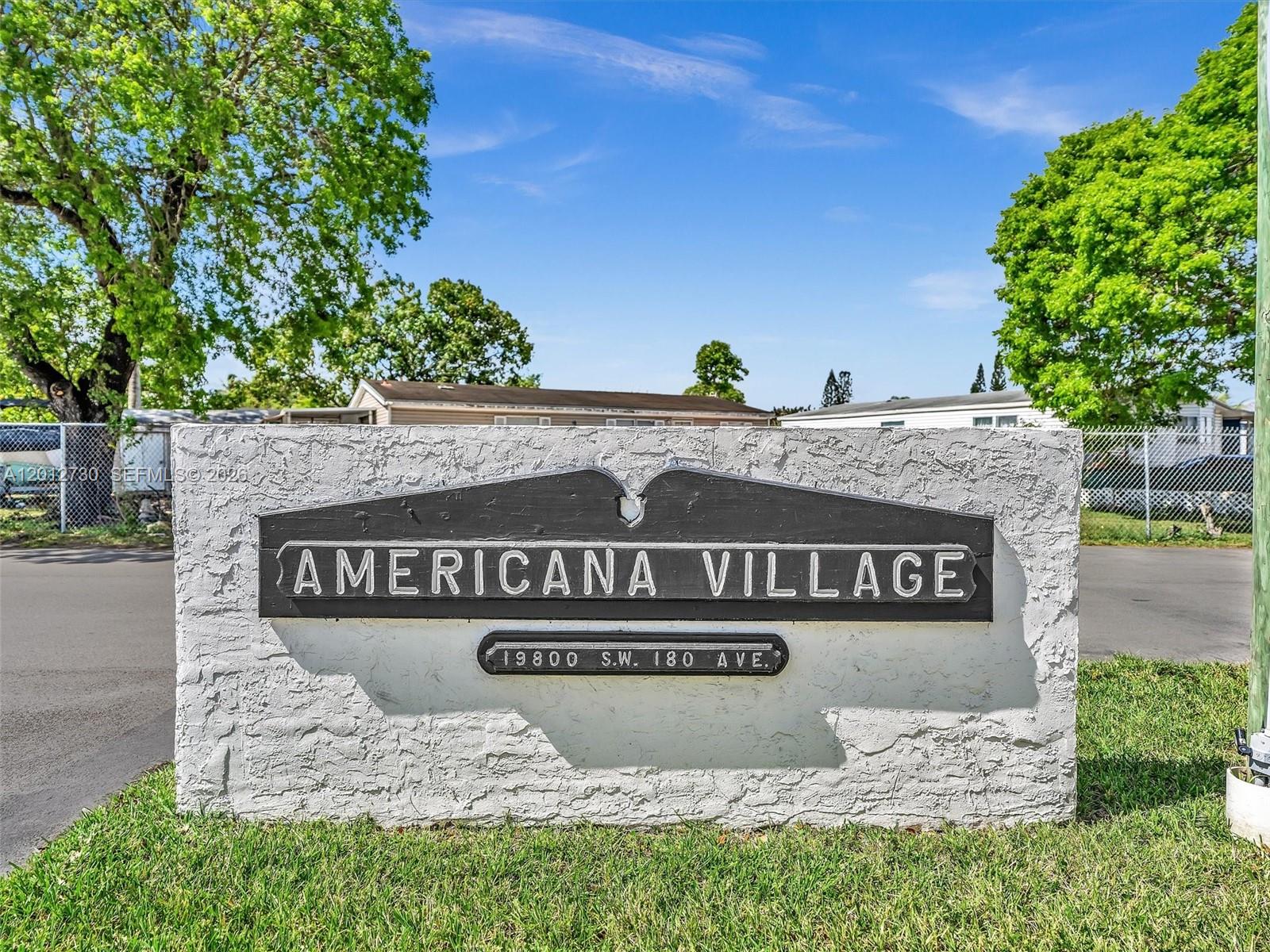 A concrete sign for Americana Village stands on a grassy lawn under a blue sky — 19800 SW 180th Ave #80, Miami FL 33187, MIAMI, FL