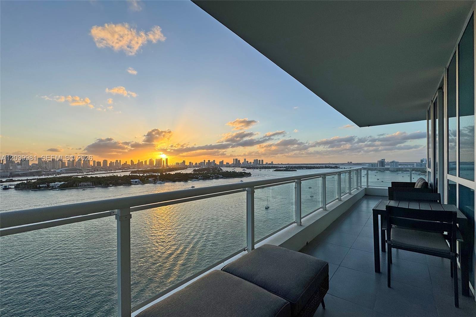A balcony with outdoor furniture overlooks a bay and a distant city skyline at sunset — 540 West Ave # 2114, Miami Beach FL 33139, MIAMI BEACH, FL