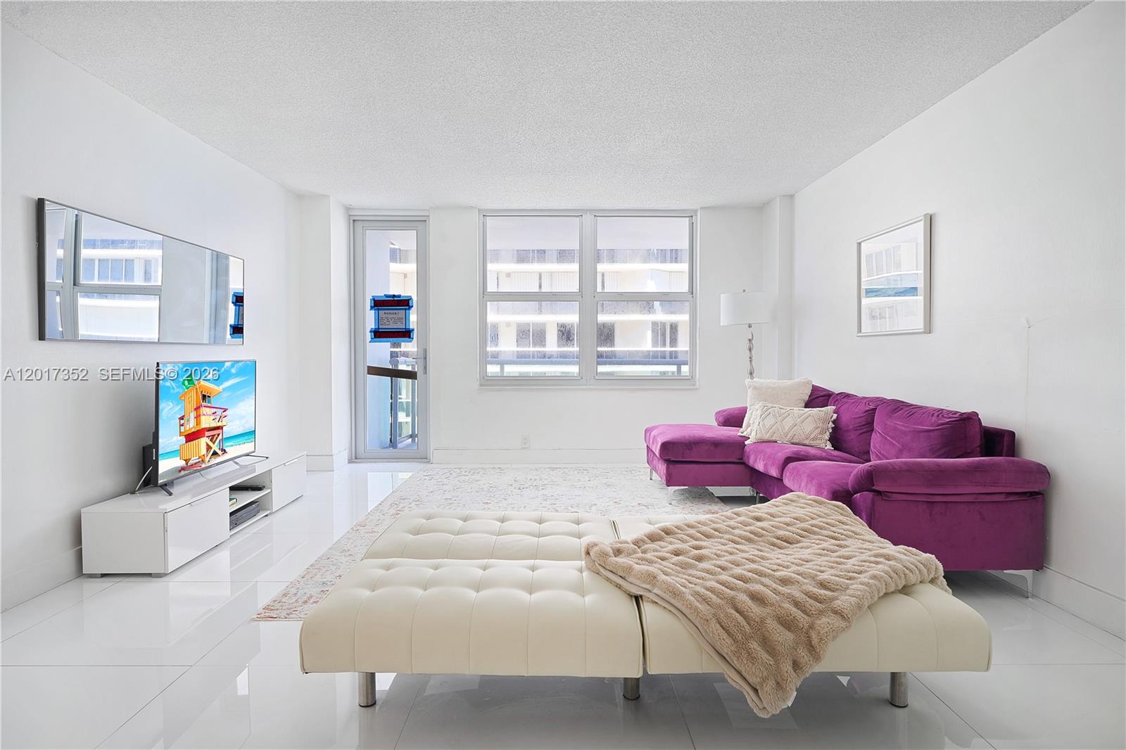 A living room features a purple sectional sofa, a white tufted ottoman, and white tile flooring — 9511 Collins Ave # 901, Surfside FL 33154, SURFSIDE, FL