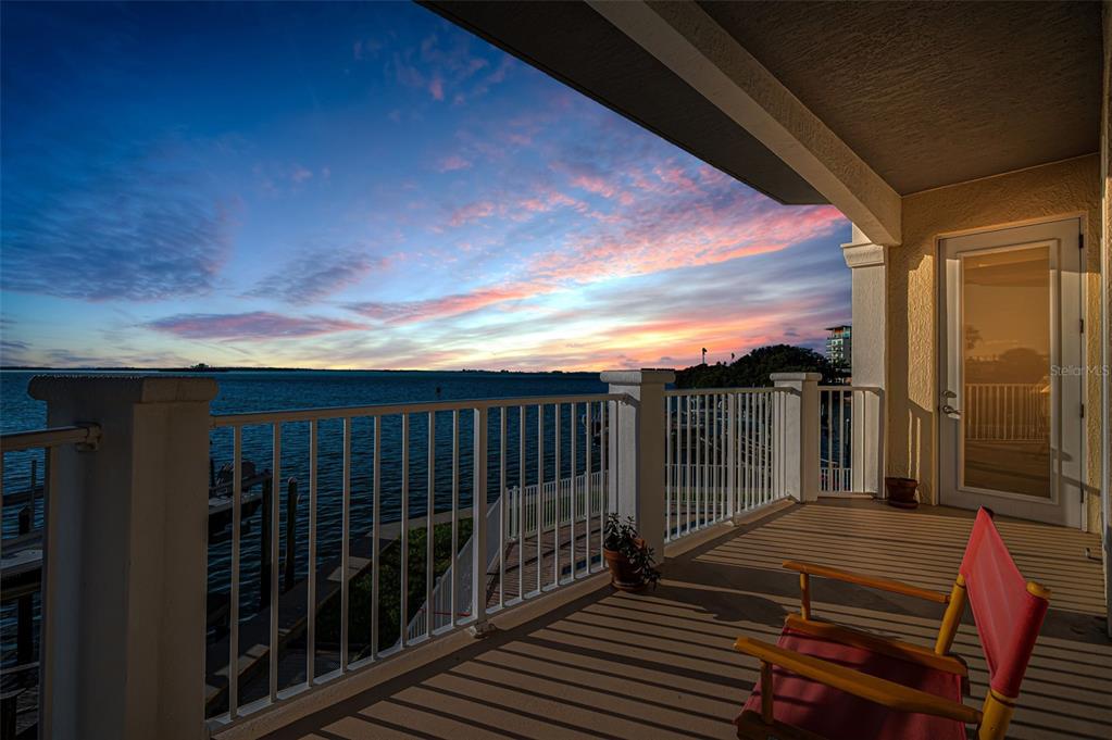A covered balcony overlooks a body of water at sunset with a red chair nearby — 1860 N FORT HARRISON AVENUE UNIT 103, CLEARWATER, FL