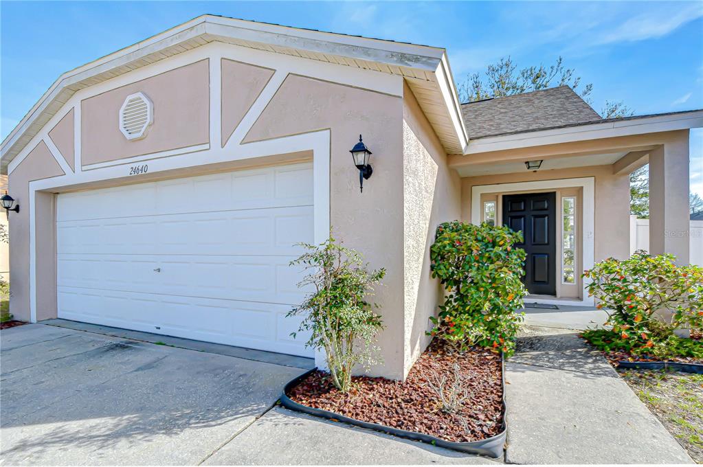 A single-story house exterior features a two-car garage, a covered entryway, and landscaped garden beds — 24640 SIENA DRIVE, LUTZ, FL