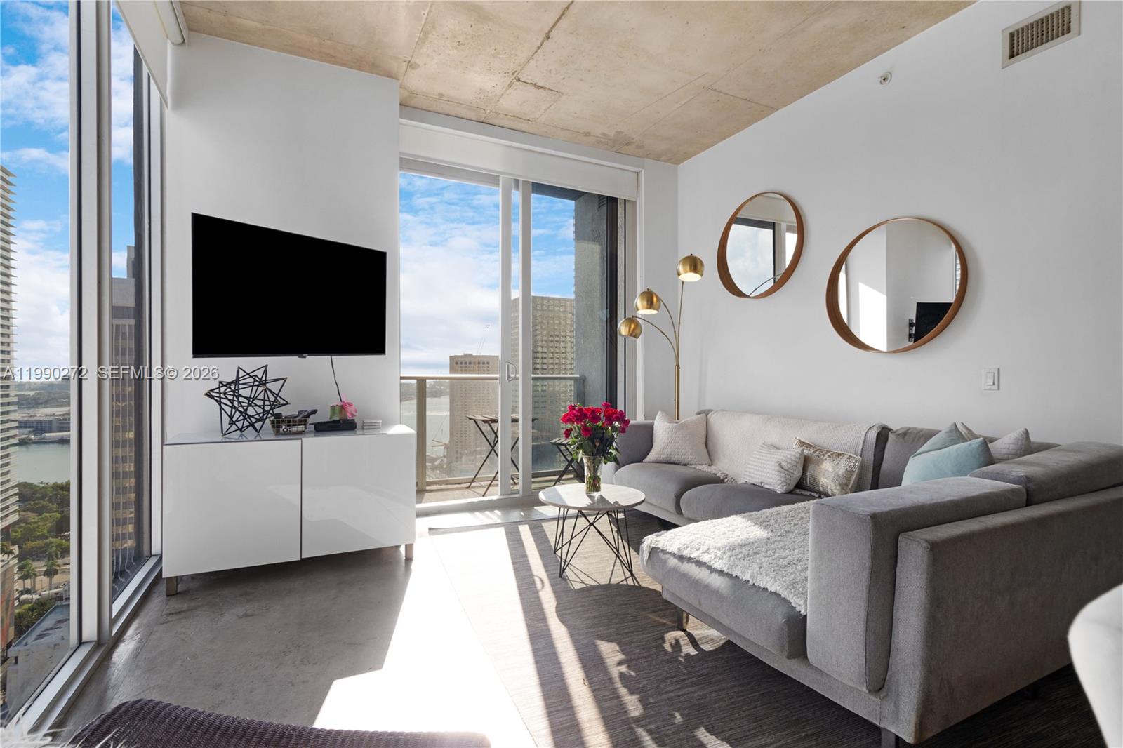 A modern living room features a gray sectional sofa, concrete ceiling, and floor-to-ceiling windows — 151 SE 1st St # 2802, Miami FL 33131, MIAMI, FL