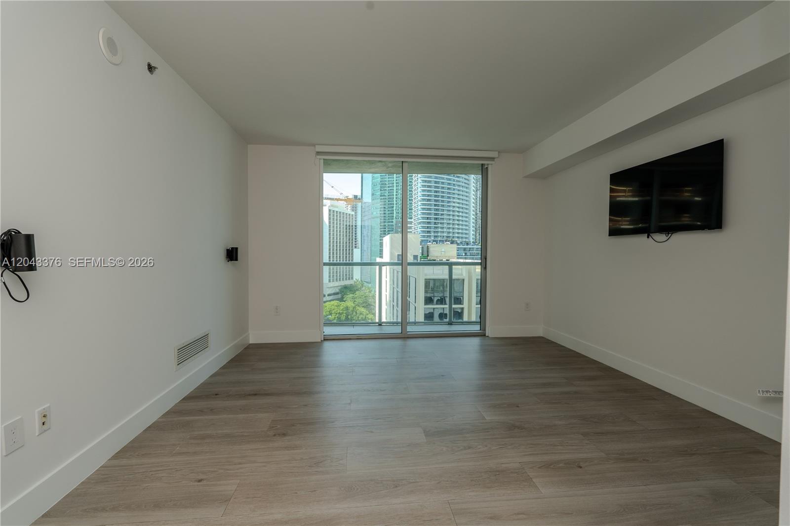 An empty room with light wood-look flooring, white walls, a wall-mounted television, and balcony access — 500 Brickell Ave # 1407, Miami FL 33131, MIAMI, FL