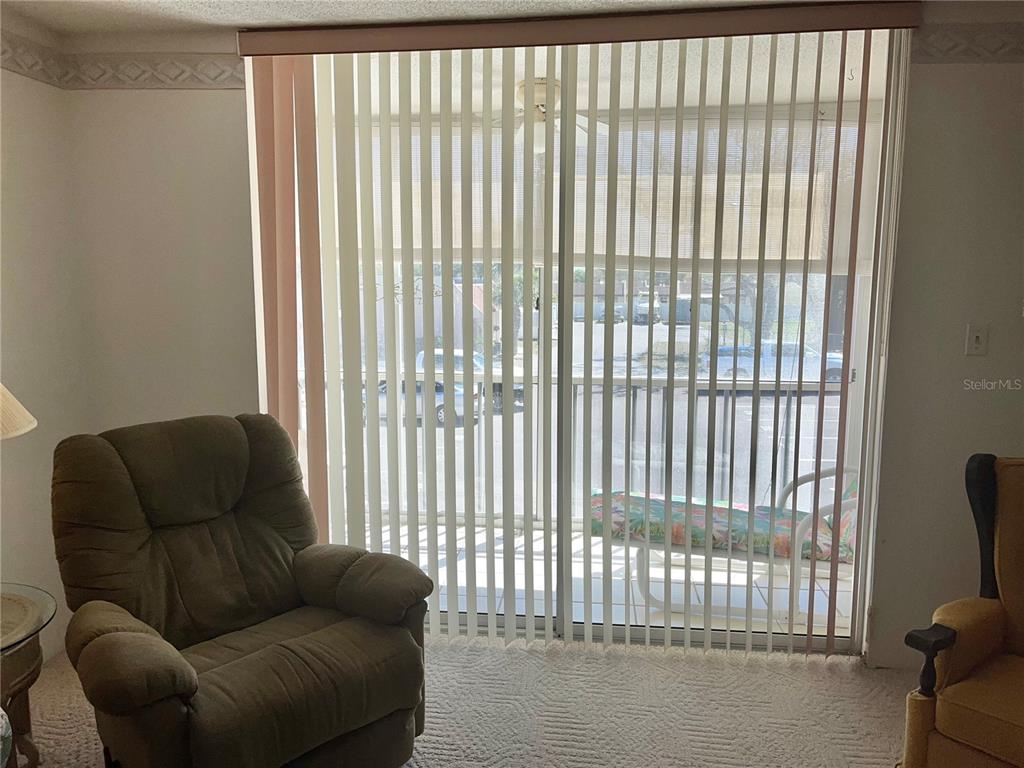 A living room features a brown recliner, vertical blinds, and a sliding glass door — 519 ALBEE FARM ROAD UNIT 210, VENICE, FL