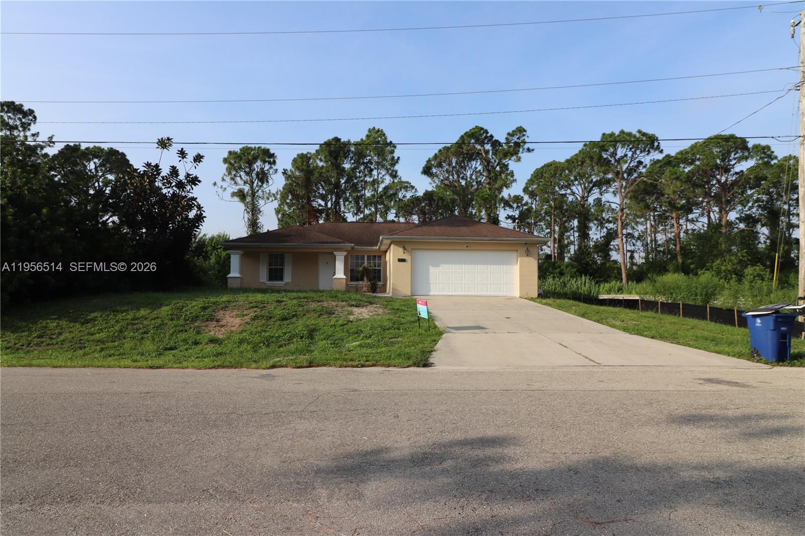 A single-story tan house with a two-car garage sits behind a sloped grassy lawn — 2805 1 sw st, Lehigh Acres FL 33972, LEHIGH ACRES, FL