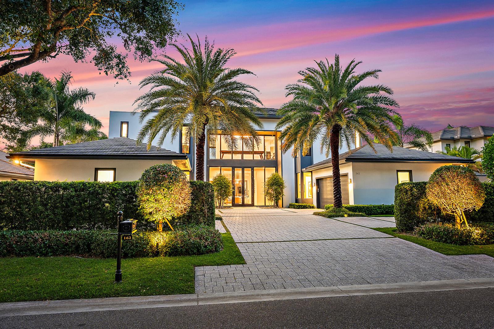A modern two-story house with a paved driveway, palm trees, and manicured hedges at dusk — 162 Commodore Drive, Jupiter FL 33477, JUPITER, FL