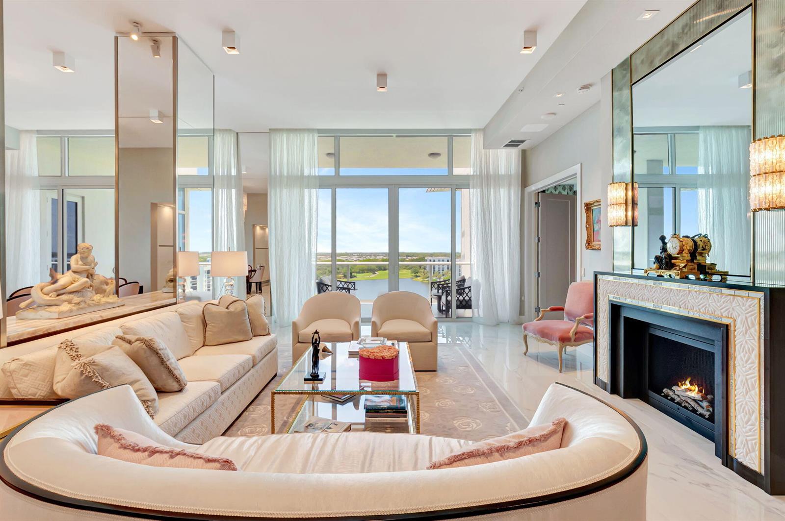 A bright living room features a fireplace, a curved sofa, and floor-to-ceiling windows overlooking water — 20155 Boca West Drive # c902, Boca Raton FL 33434, BOCA RATON, FL
