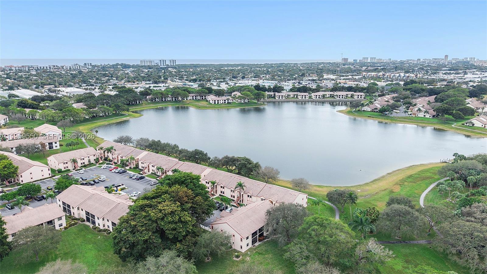 An aerial view shows multi-story residential buildings surrounding a large lake in a suburban area — 3150 Leewood Ter # L124, Boca Raton FL 33431, BOCA RATON, FL