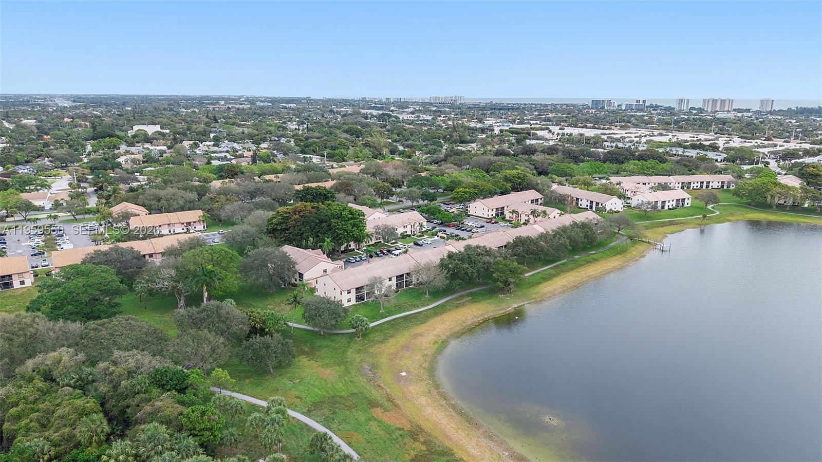 An aerial view shows multi-story residential buildings situated next to a large lake and trees — 3150 Leewood Ter # L124, Boca Raton FL 33431, BOCA RATON, FL