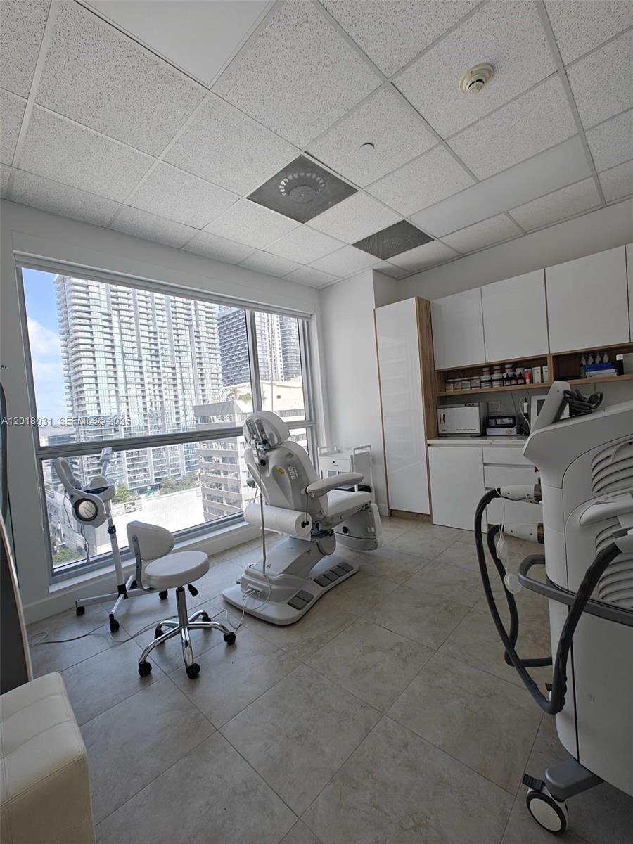 A medical treatment room features a procedure chair, rolling stool, equipment, and city skyline views — 175 SW 7th St # 2006, Miami FL 33130, MIAMI, FL