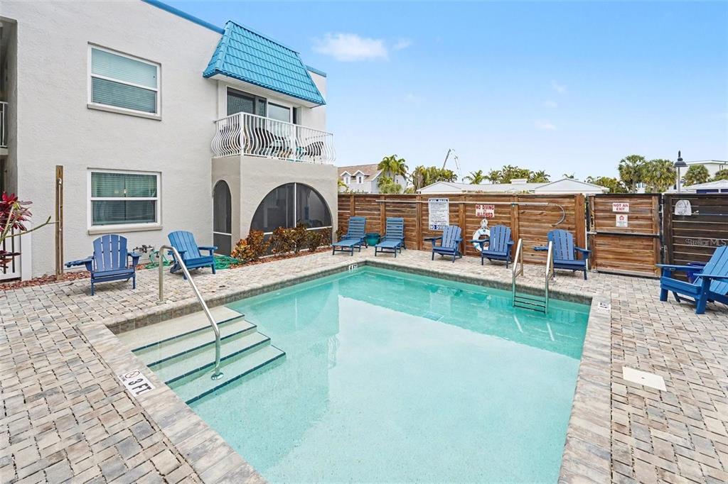 A swimming pool with blue lounge chairs sits on a brick patio beside a building — 17035 GULF BOULEVARD UNIT 216, ST PETERSBURG, FL