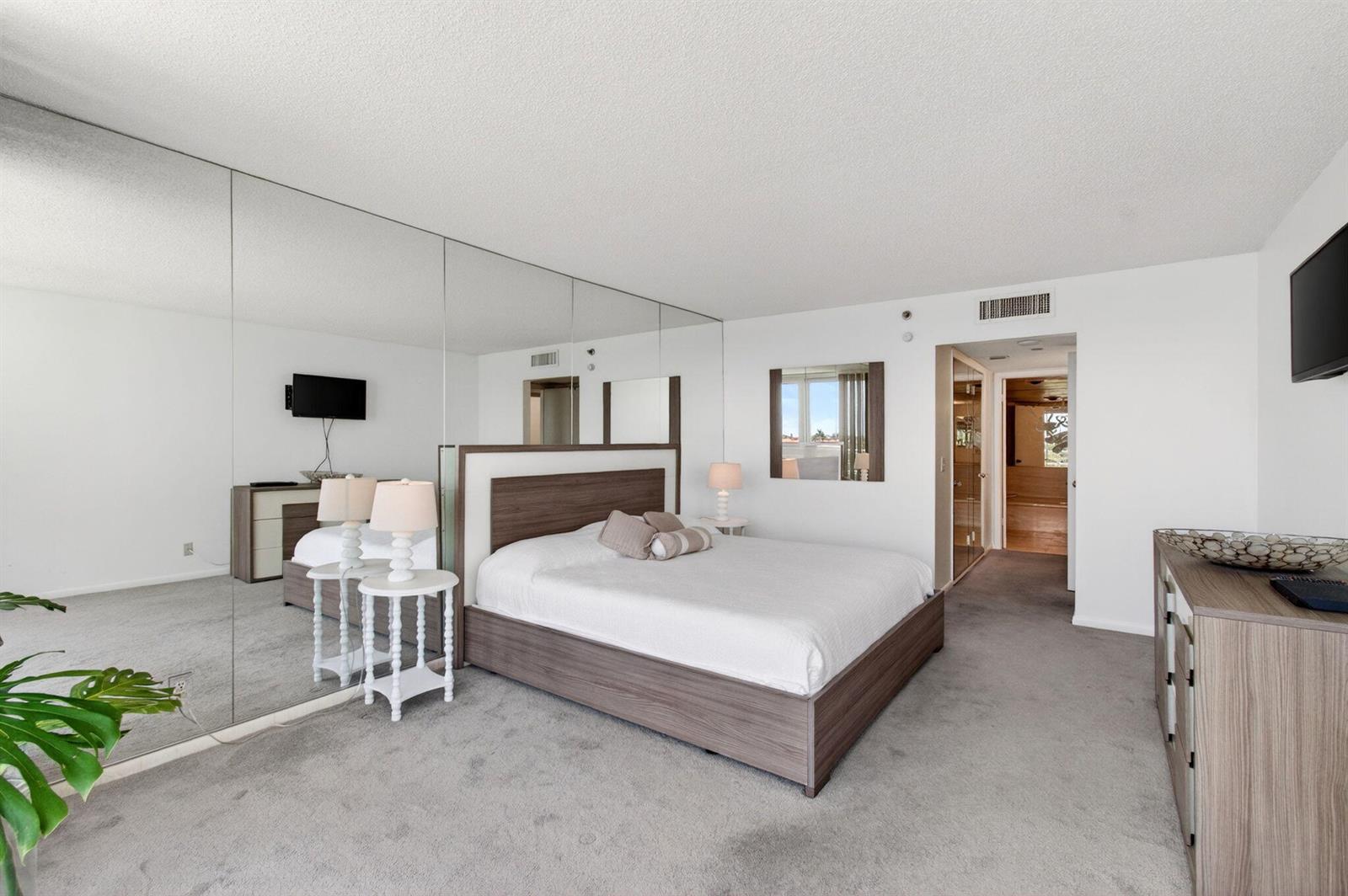 A bedroom features a wood-frame bed, mirrored wall, gray carpeting, and white walls — 4201 N Ocean Boulevard # 307, Boca Raton FL 33431, BOCA RATON, FL