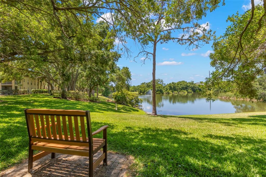A wooden bench sits on a brick patio overlooking a grassy lawn and a pond — 2690 CORAL LANDINGS BOULEVARD UNIT 112, PALM HARBOR, FL