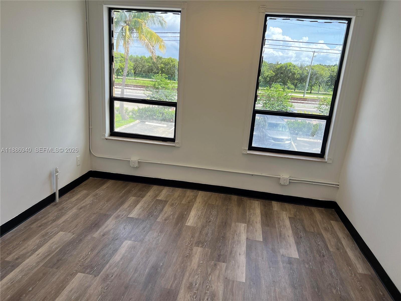 An empty room features wood-look flooring, white walls, and two windows overlooking a parking lot — 2001 N Dixie Hwy, Pompano Beach FL 33060, POMPANO BEACH, FL