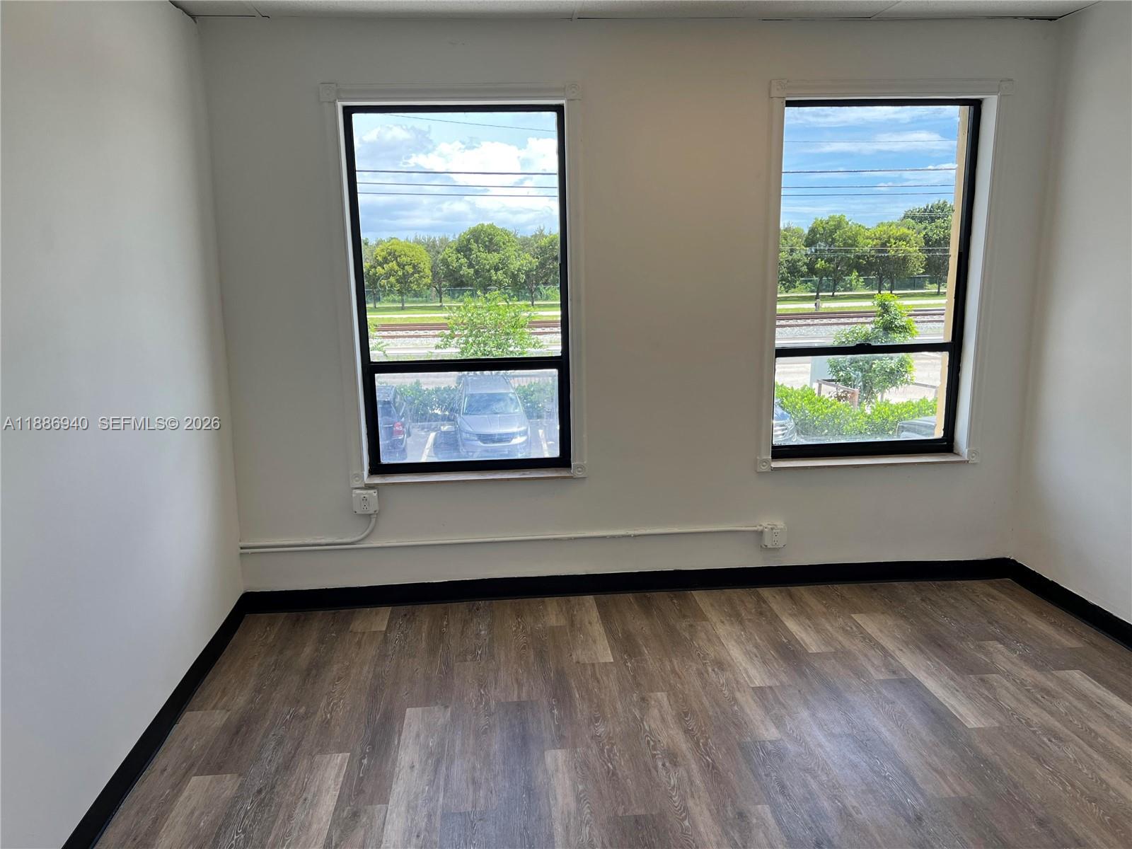 An empty room features wood-look flooring, white walls, and two windows overlooking a parking lot — 2001 N Dixie Hwy, Pompano Beach FL 33060, POMPANO BEACH, FL