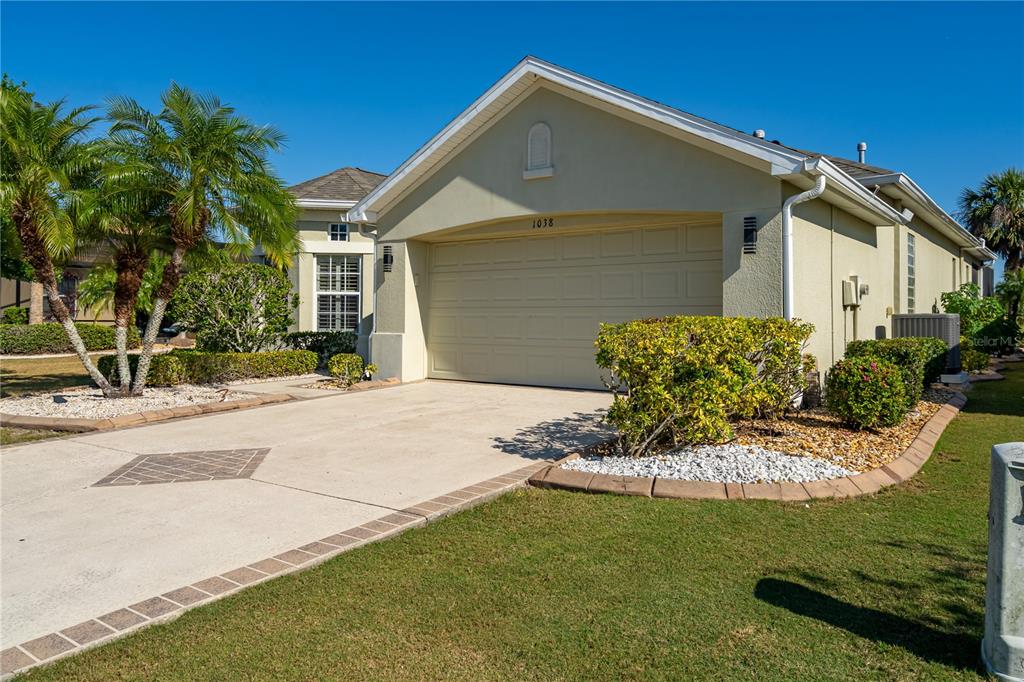 A single-story house exterior features a beige stucco facade, a two-car garage, and landscaped lawn — 1038 EMERALD DUNES DRIVE, SUN CITY CENTER, FL