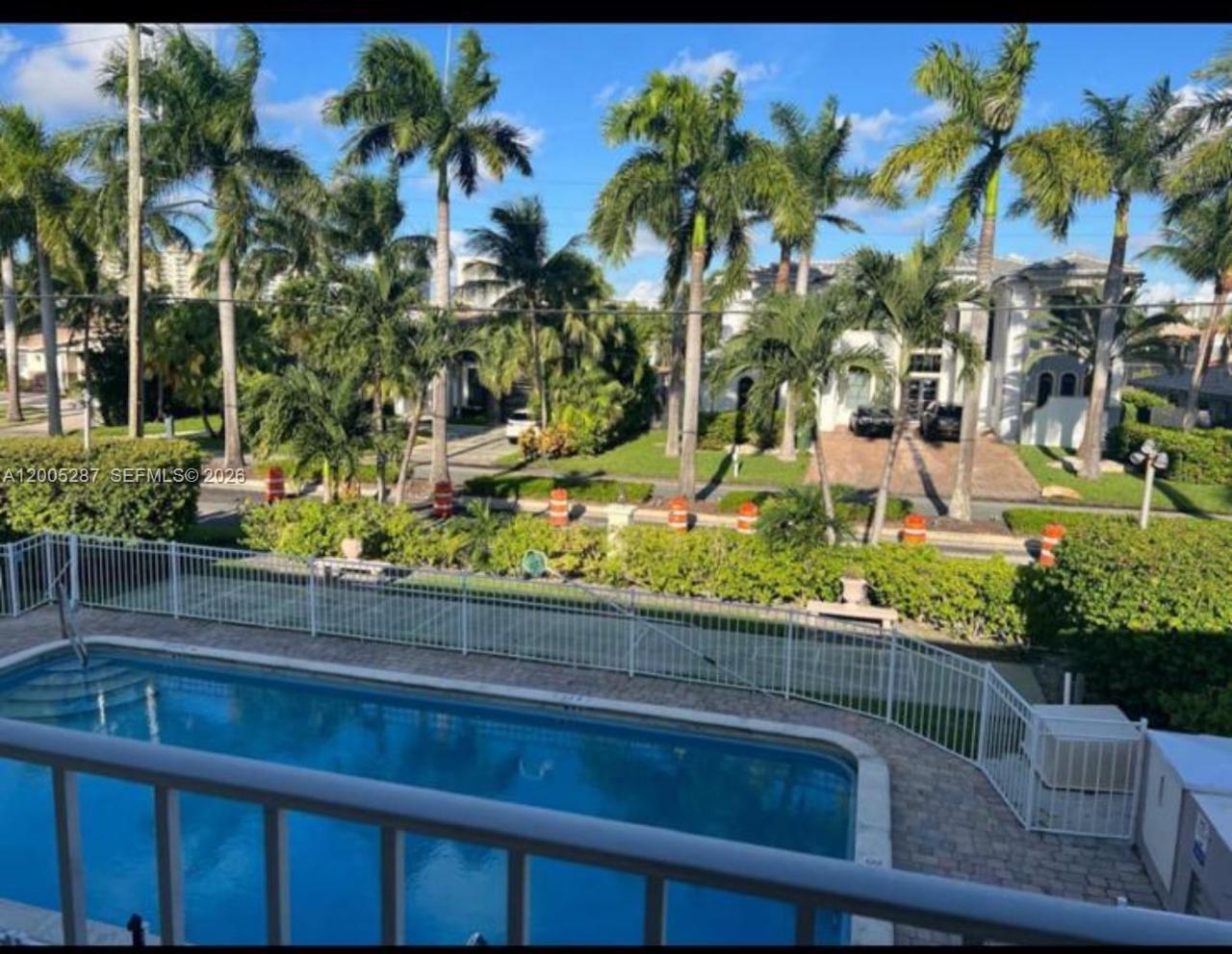 An elevated view of a swimming pool with a white fence and palm trees beyond — 500 Layne Blvd # 14, Hallandale Beach FL 33009, HALLANDALE BEACH, FL