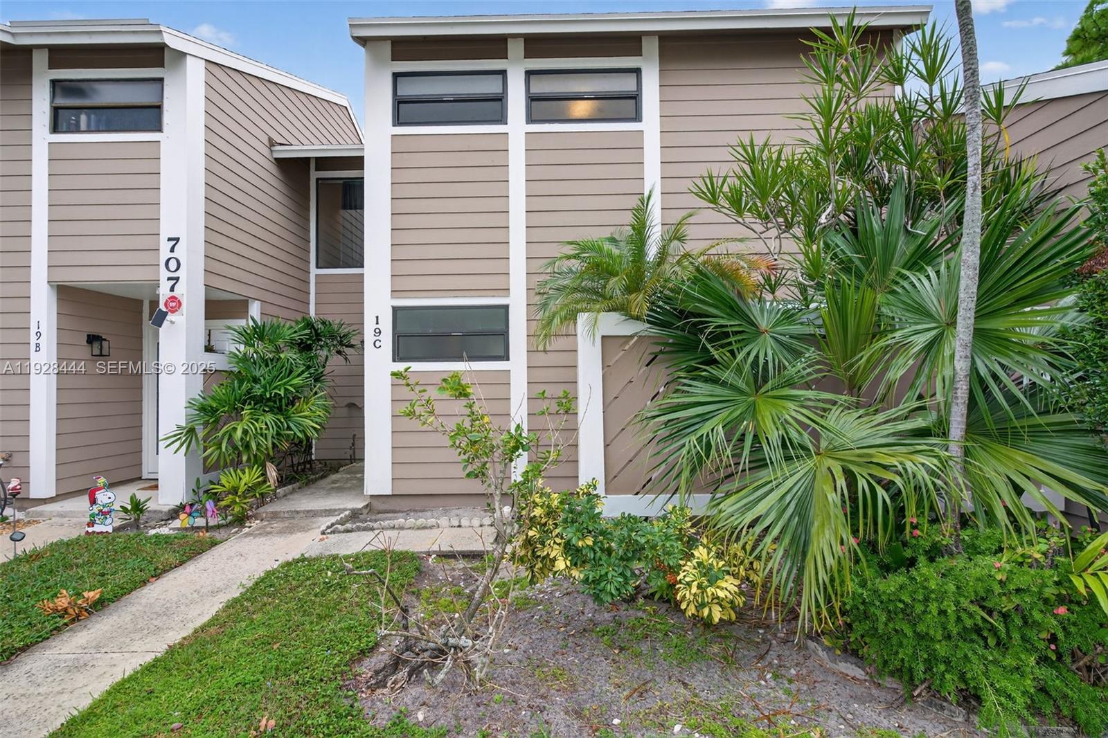 The exterior of a two-story townhouse features tan horizontal siding, white trim, and lush landscaping — 707 Stonewood Ct # 19C, Jupiter FL 33458, JUPITER, FL