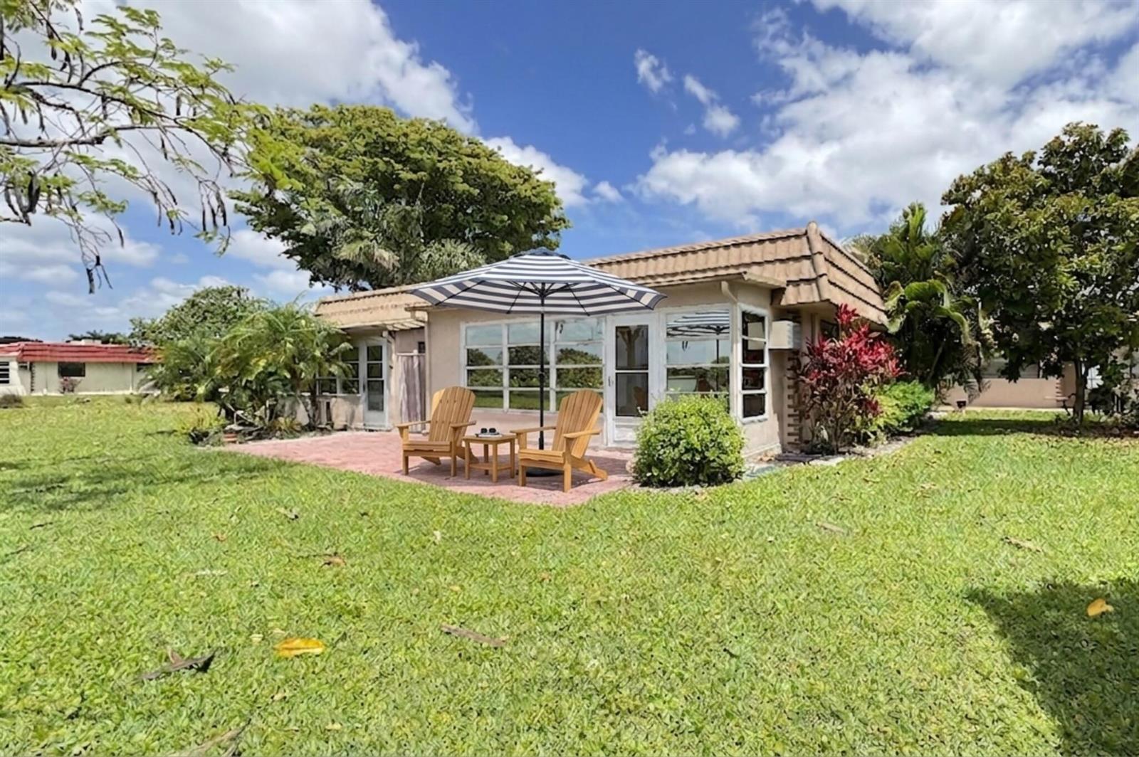 A single-story house with a patio, two wooden chairs, and a striped umbrella on grass — 220 Seville J, Delray Beach FL 33446, DELRAY BEACH, FL