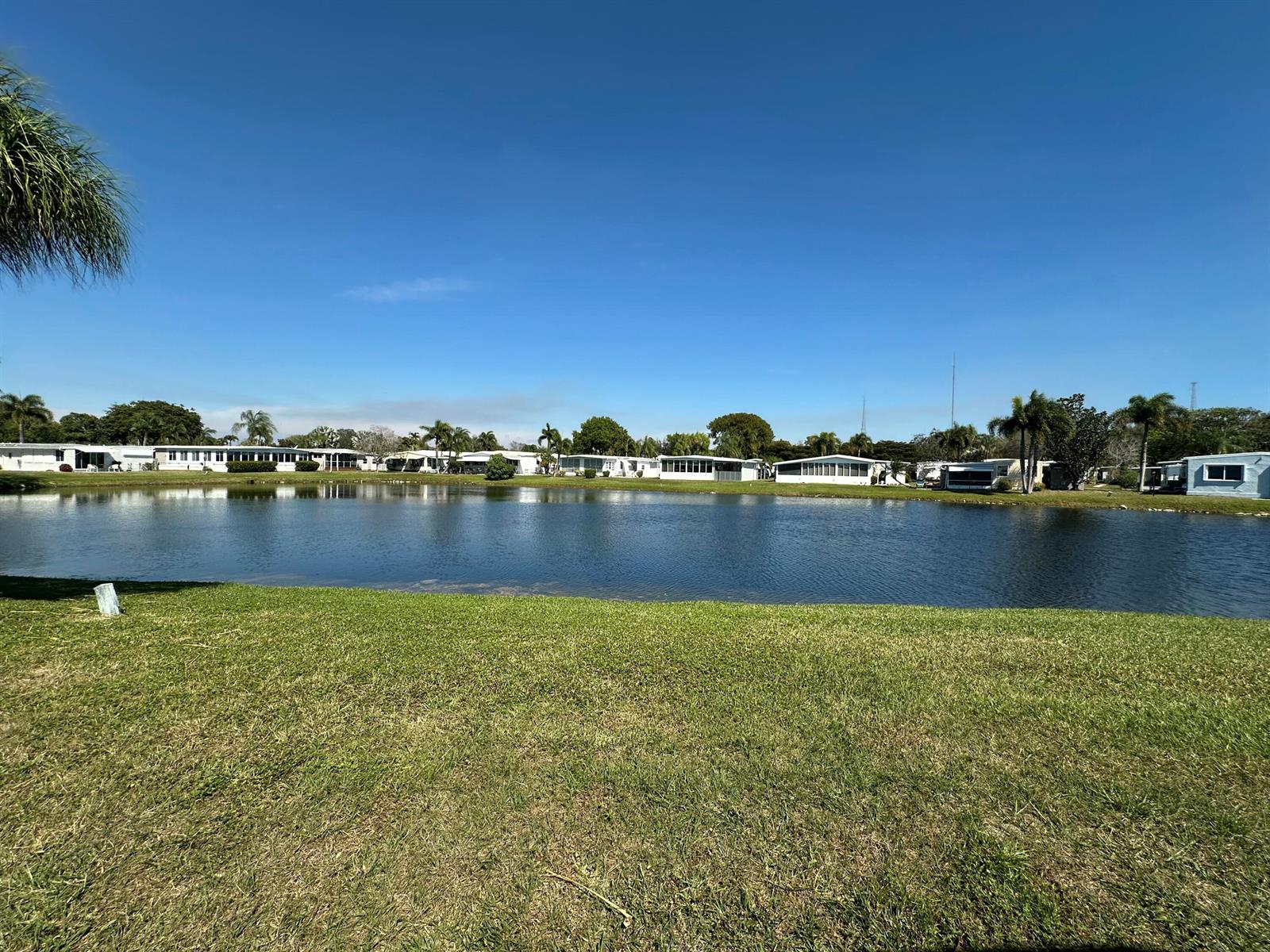 A grassy lawn overlooks a pond with a row of mobile homes in the background — 8349 South Street, Boca Raton FL 33433, BOCA RATON, FL