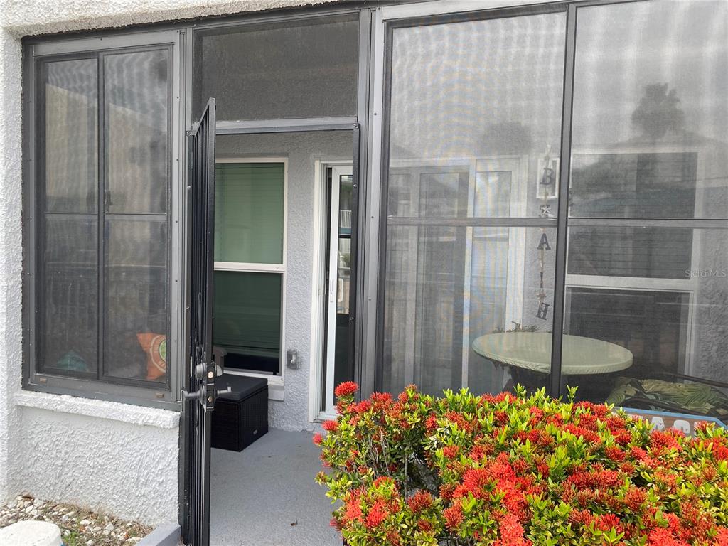 A screened-in patio features a black metal door, a small table, and red flowering shrubs — 230 MEDALLION BOULEVARD UNIT D, MADEIRA BEACH, FL