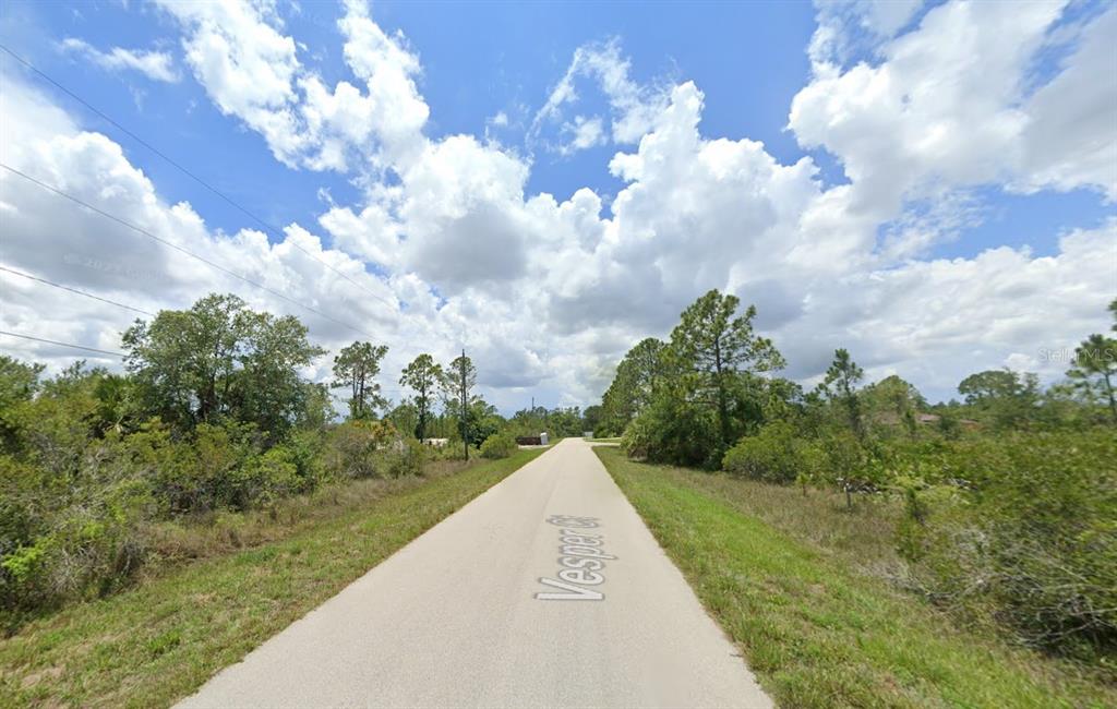 A paved road stretches through a wooded area under a bright, cloudy sky — 1928 VESPER COURT, LEHIGH ACRES, FL
