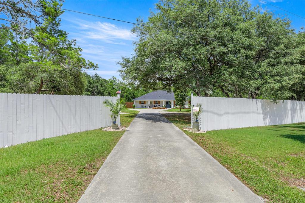 A concrete driveway leads through a white fence toward a house shaded by large trees — 495 FISHER ROAD, OCKLAWAHA, FL