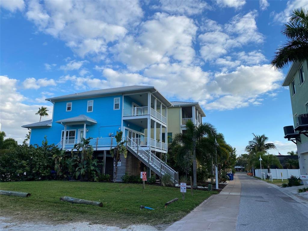 A bright blue elevated house with a white staircase and porch stands beside a paved road — WHISPERING PINES DRIVE, INDIAN SHORES, FL