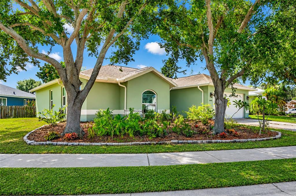 A single-story green house with a front garden, two large trees, and a driveway — 2405 ICECAPADE DRIVE, SARASOTA, FL